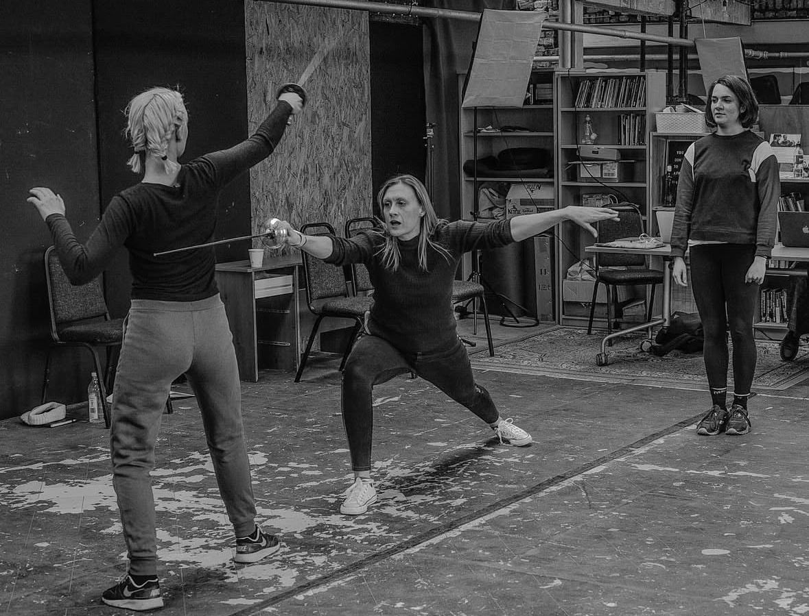 black and white photo of 3 women, the middle woman is fighting with a sword and lunging forward as if fencing.