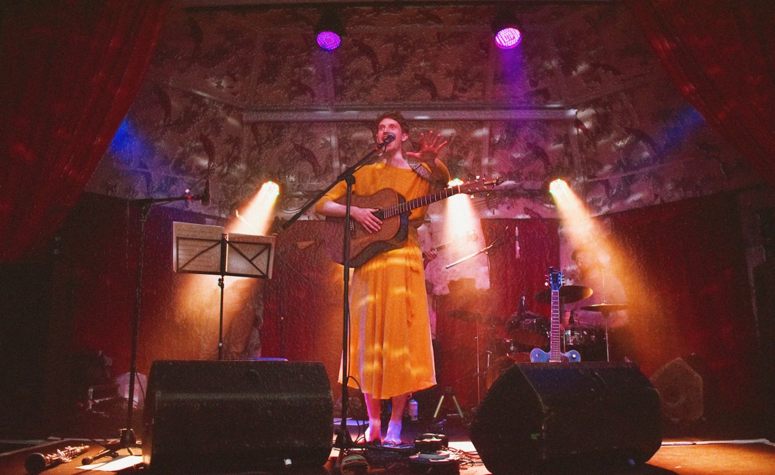 Bay Bryan centre stage. They are wearing a yellow dress and holding an acoustic guitar.