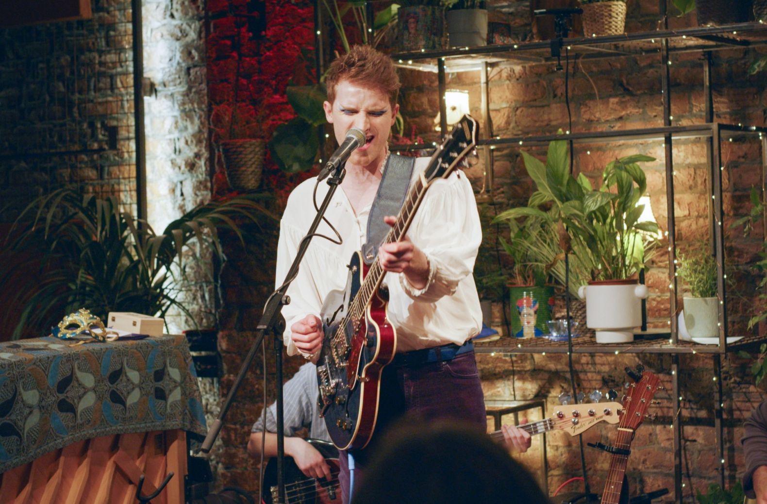 Bay Bryan, dressed in blue jeans and a white shirt, is playing a guitar and singing into a microphone. There is another musician seated behind, partially obscured by Bay Bryan.