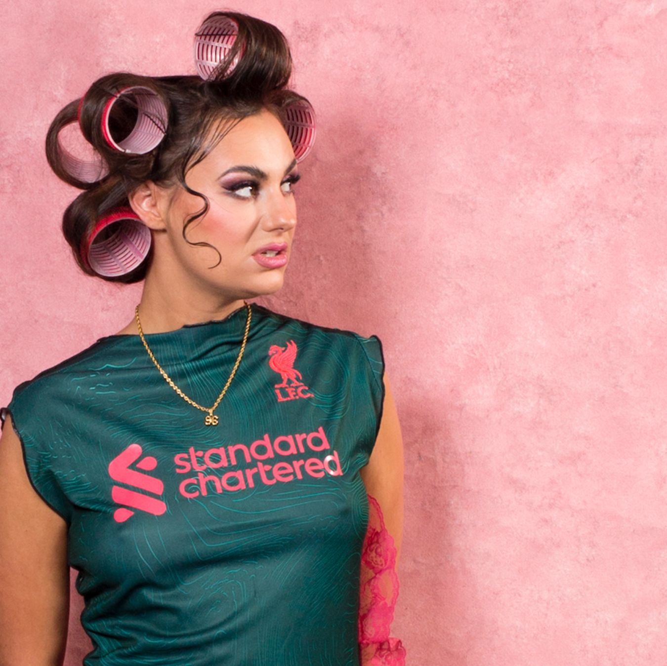 woman with hair in rollers stood in front of a pink background wearing a Liverpool FC shirt