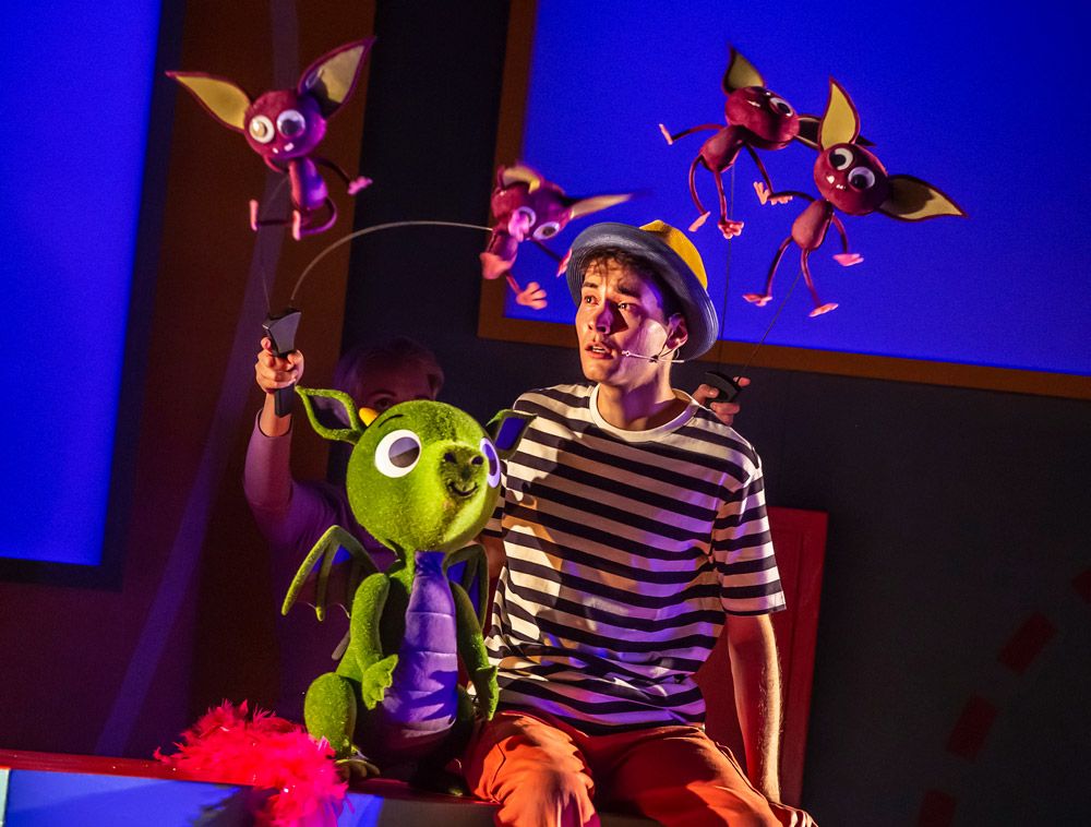 An actor sitting with a green dragon puppet and smaller bat-like puppets flying overhead.