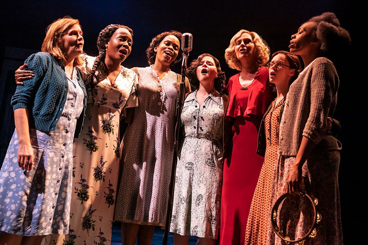 seven ladies around an old fashioned microphone, singing