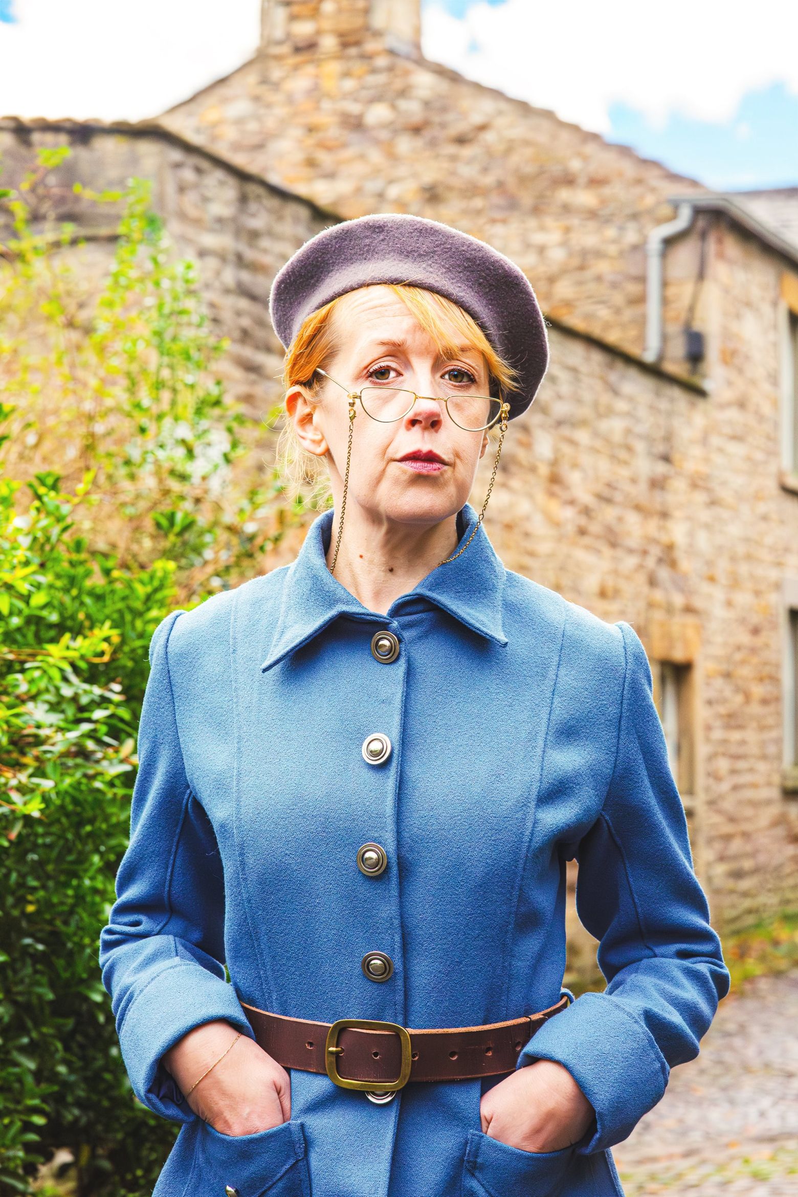 Eslie Orr stood front of stone house, she is wearing a blue suit and a blue beret.