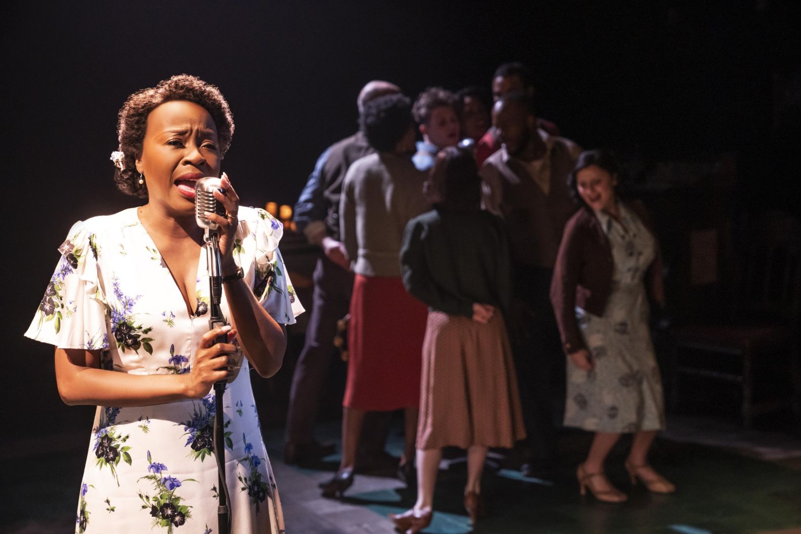 a lady singing into an old fashioned microphone with actors in the background dancing