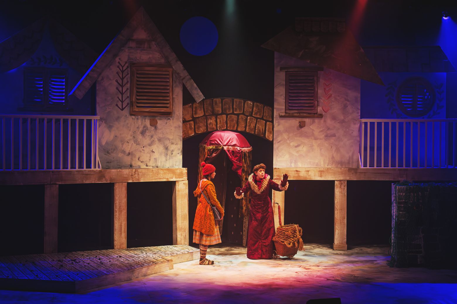 The Snow Queen at the Dukes - Gerda meets a magic woman dressed in a full length red velvet coat. they are stood centre stage infront of a set that looks like a village.