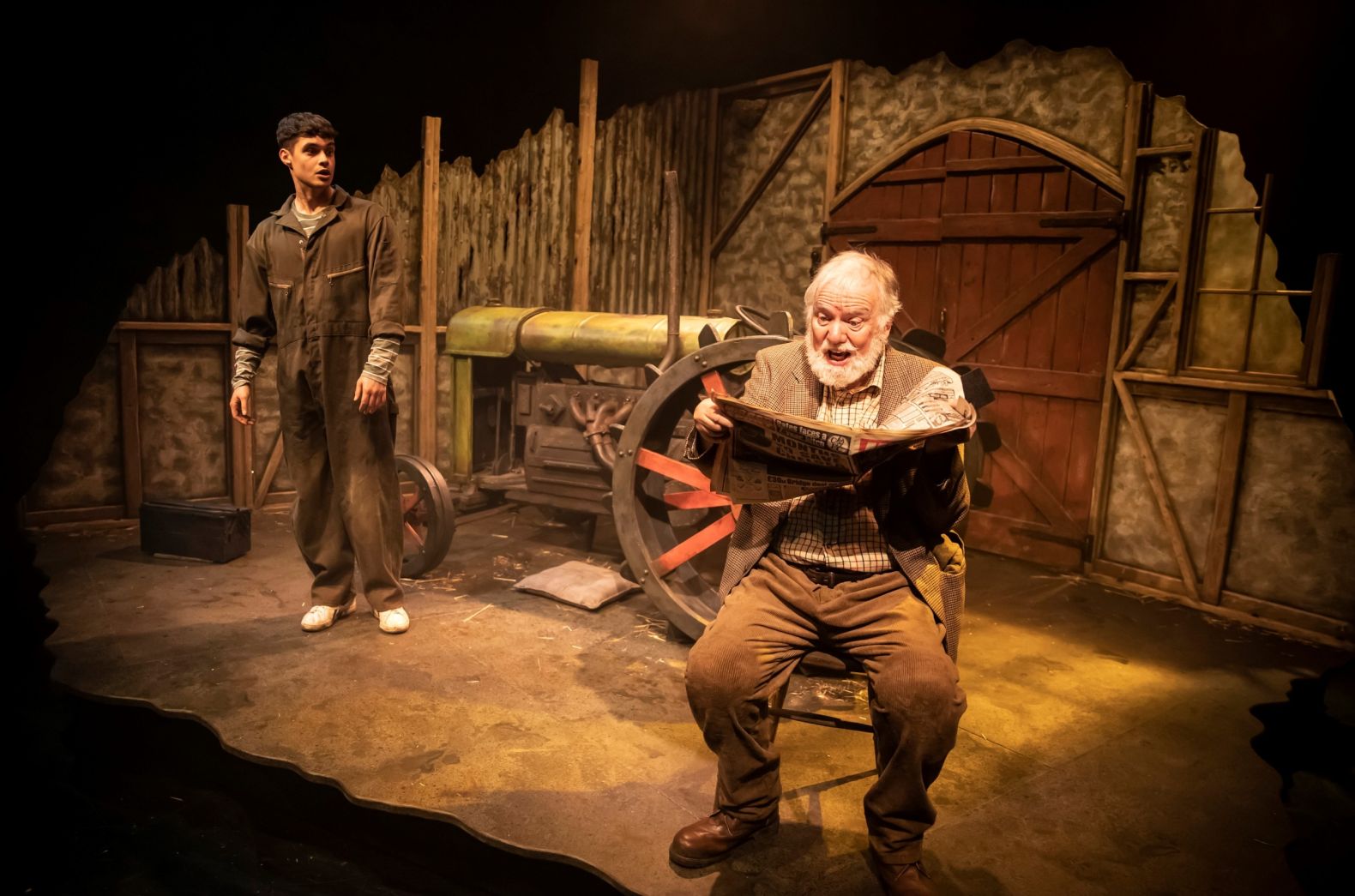 A young man and an old man in a farm setting.  The old man, in the foreground, is seated reading a newspaper.