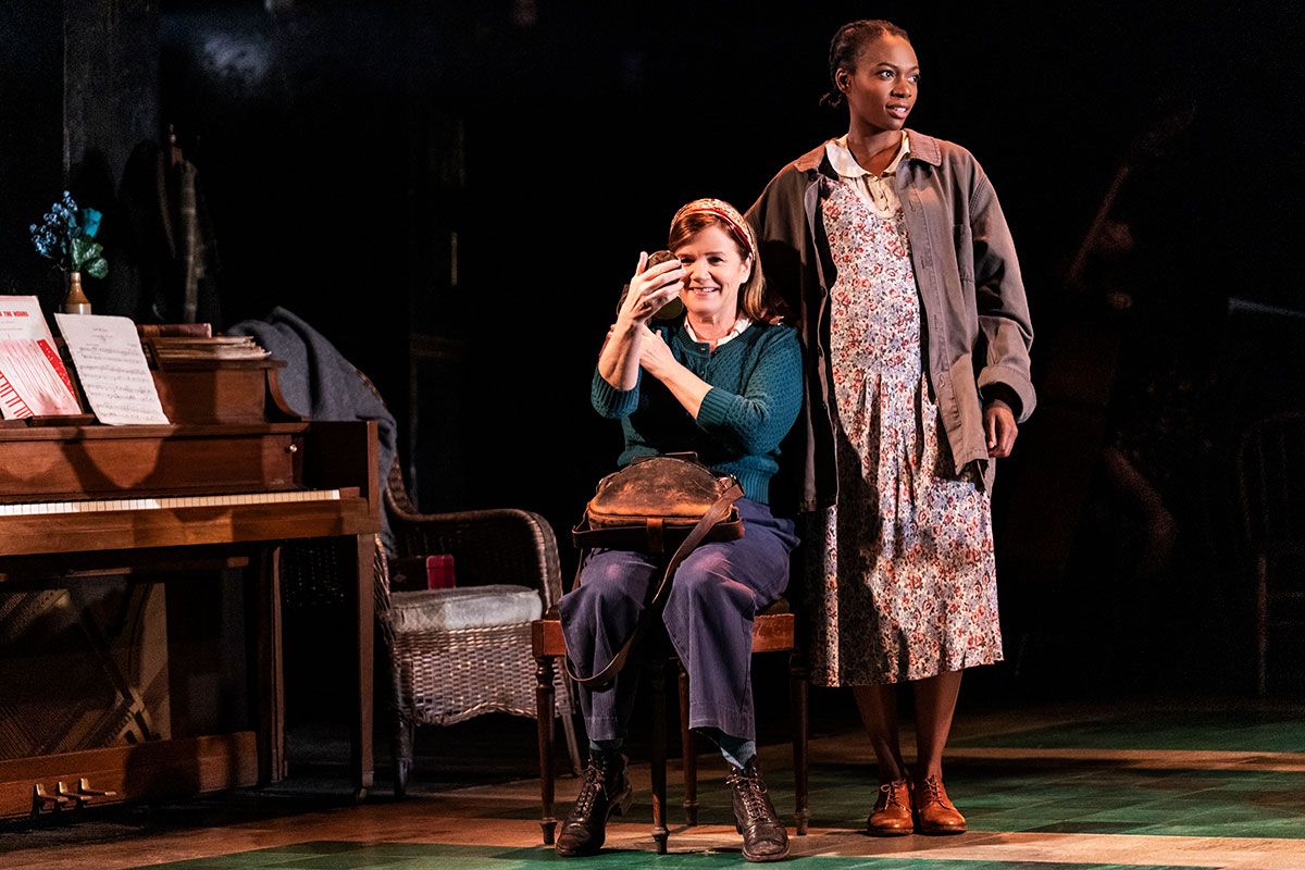 a woman sat on a chair with handbag in lap, admiring herself in a compact mirror.  Another woman stands to her left