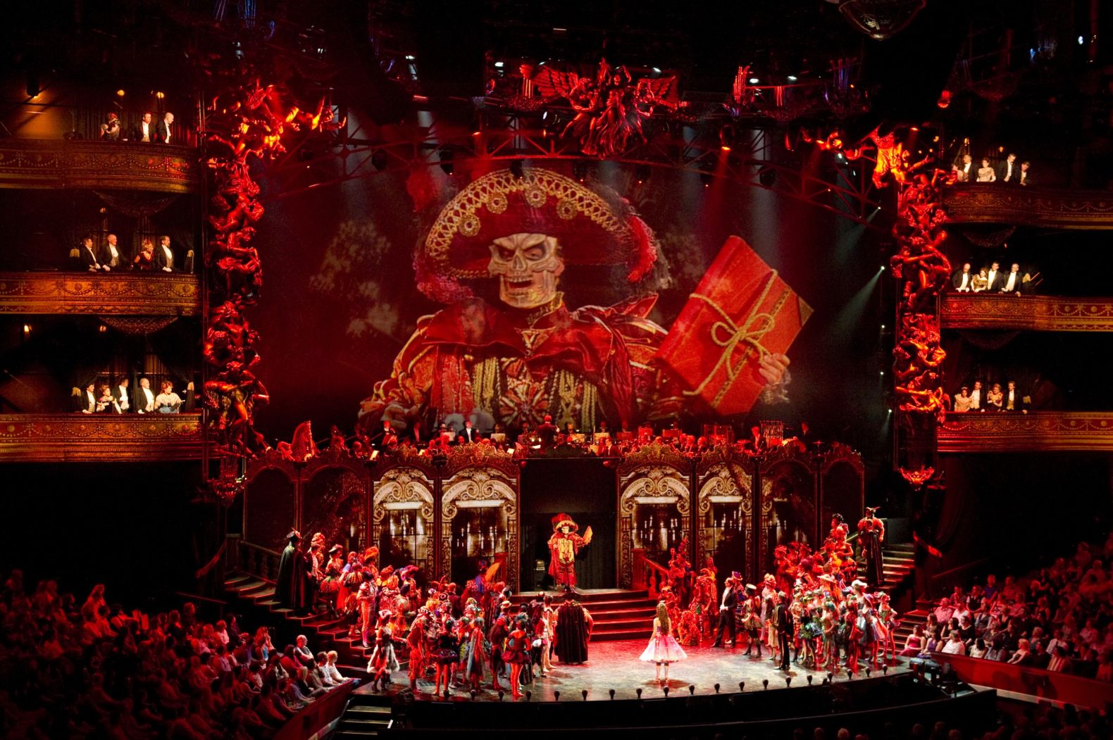 a view of the stage from the back of the auditorium.  An image of a masked individual projected above the actors