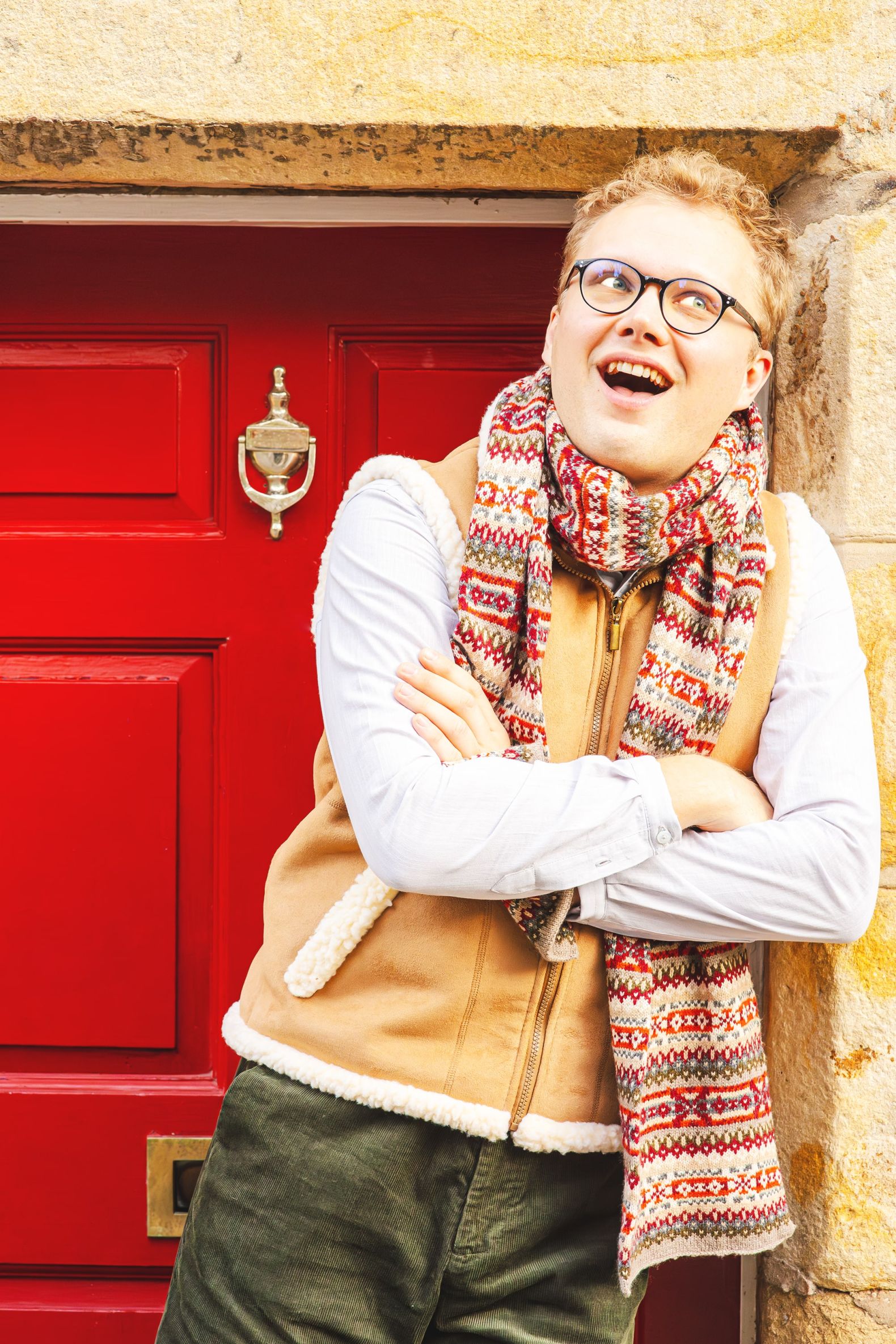 The Snow Queen at the Dukes - Kai leans against stone doorway wearing a sheep skin gilet and a red scarf.