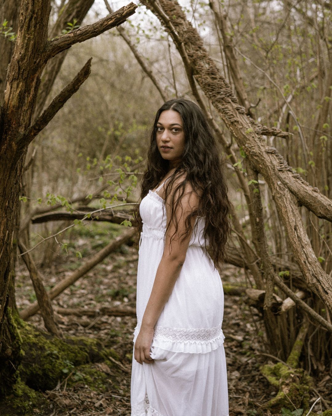 performer stood in a woodland area, they are wearing a white dress and look over their shoulder down the lens of the camera - Macbeth by HER Productions at The Dukes, Lancaster