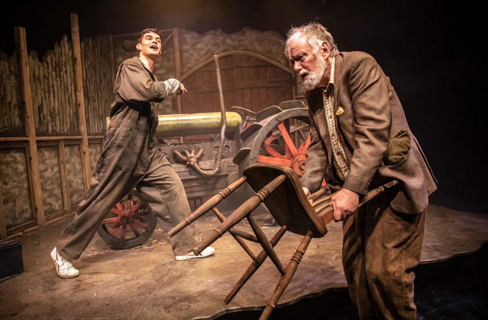 a young man and an old man in a farm setting.  The old man, in the foreground, has picked up a wooden chair and is looking weary.  