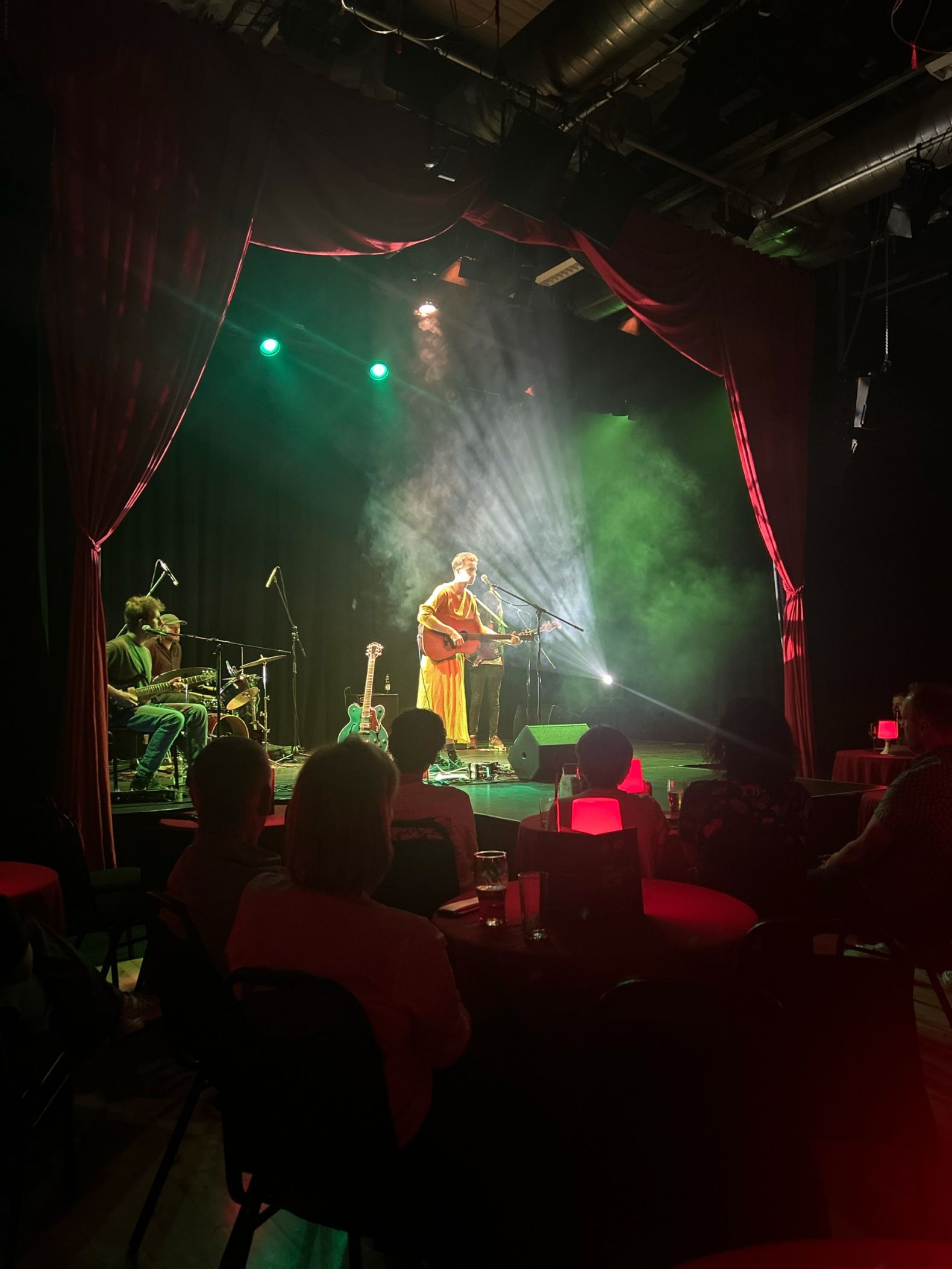 Bay Bryan performing on stage. The photograph is taken from behind the audience. Bay Brian, bathed in light, is playing a guitar and singing into a microphone.