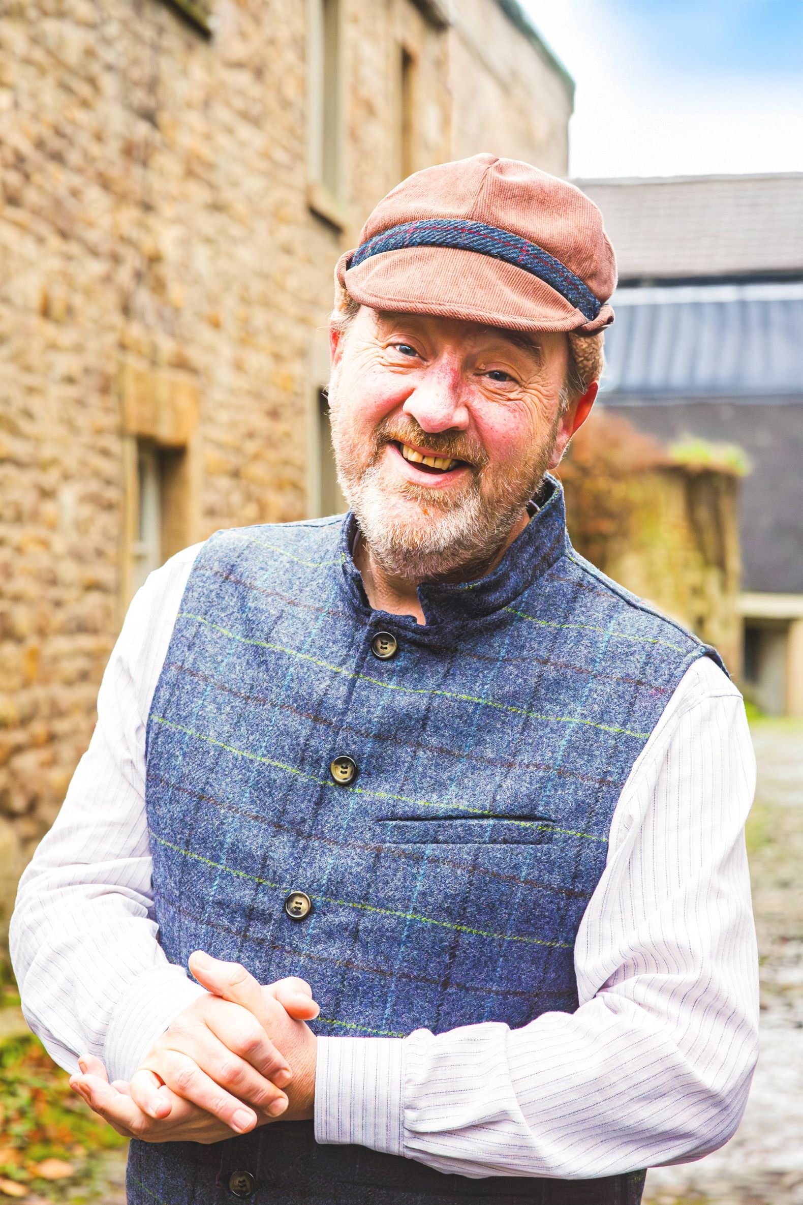 The Snow Queen at The Dukes - Mr Kelpine is looking to camera smiling, he is wearing a blue tweed waistcoat and a red deer stalker hat.