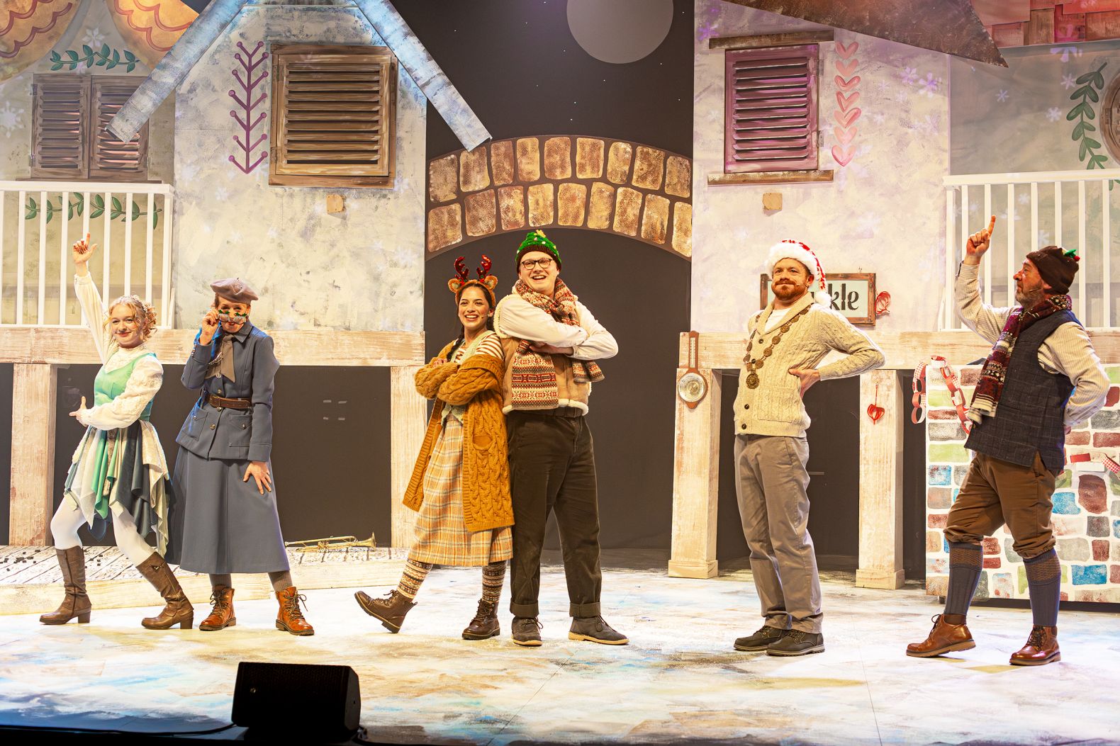 The Snow Queen at The Dukes - the 6 cast memebers of The Snow Queen are stood in various poses in a line in front of the set. The Set is a village made of houses on stilts.
