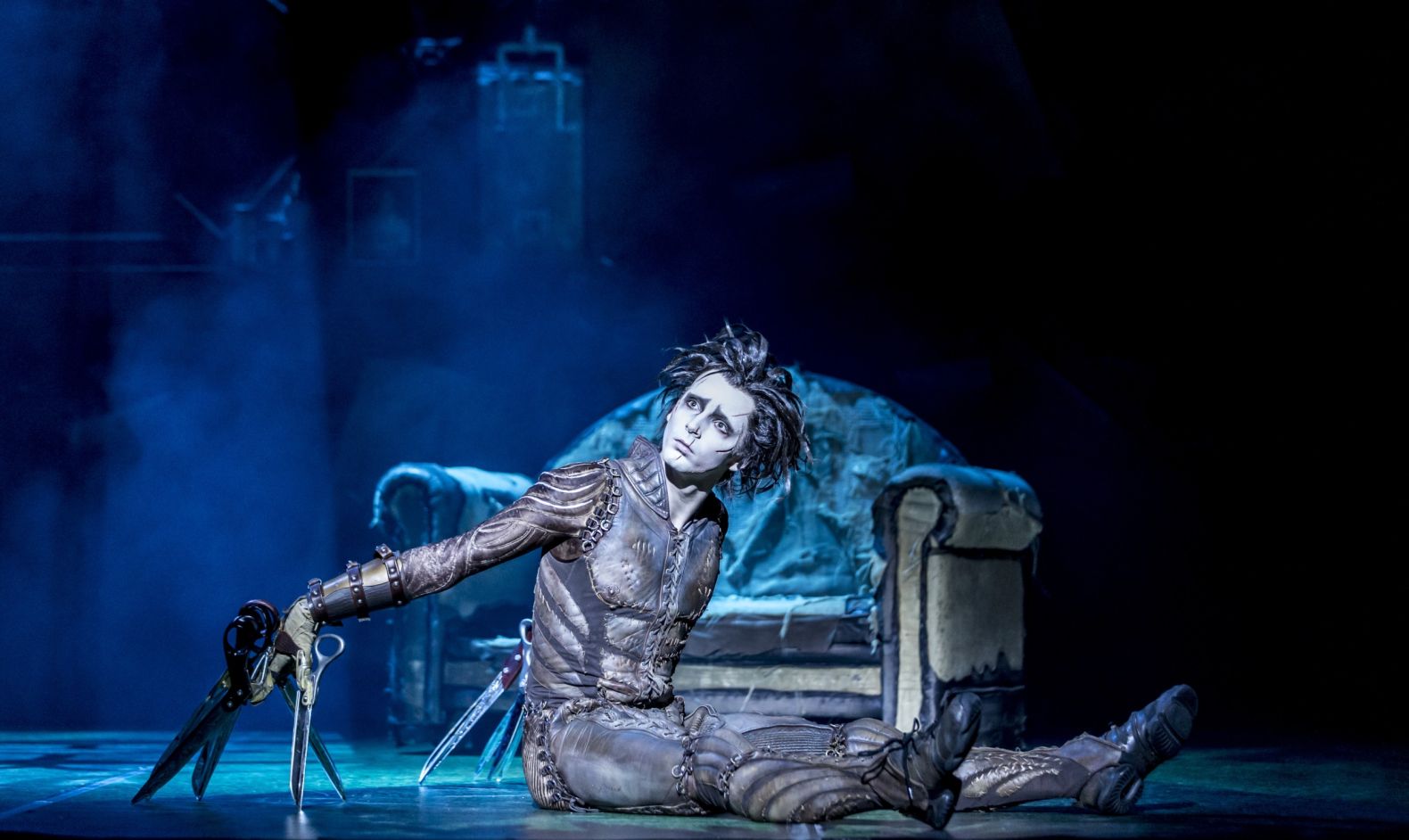 Edward sits on the floor of a dark room with mist. In the background is an old sofa. 