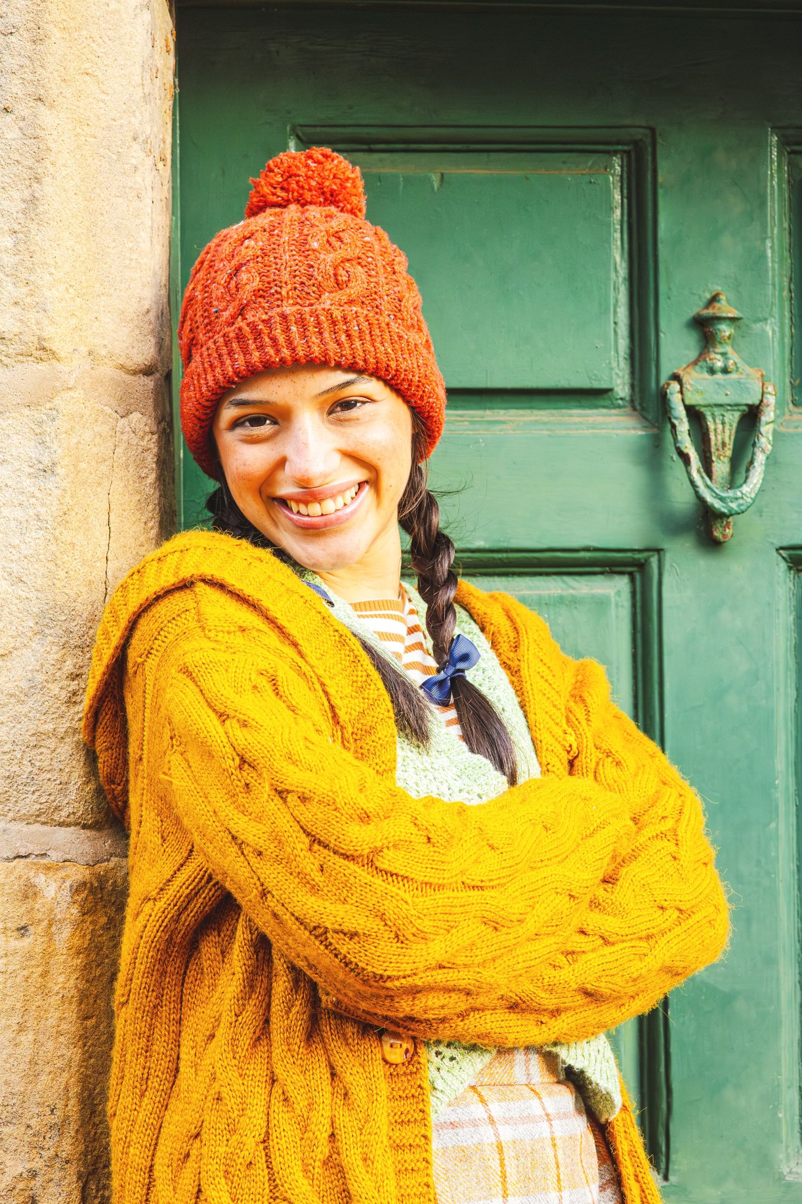 The Snow Queen at The Dukes - Gerda leans against a stone doorway wearing a chunky mustard cardgian and an orange hat.