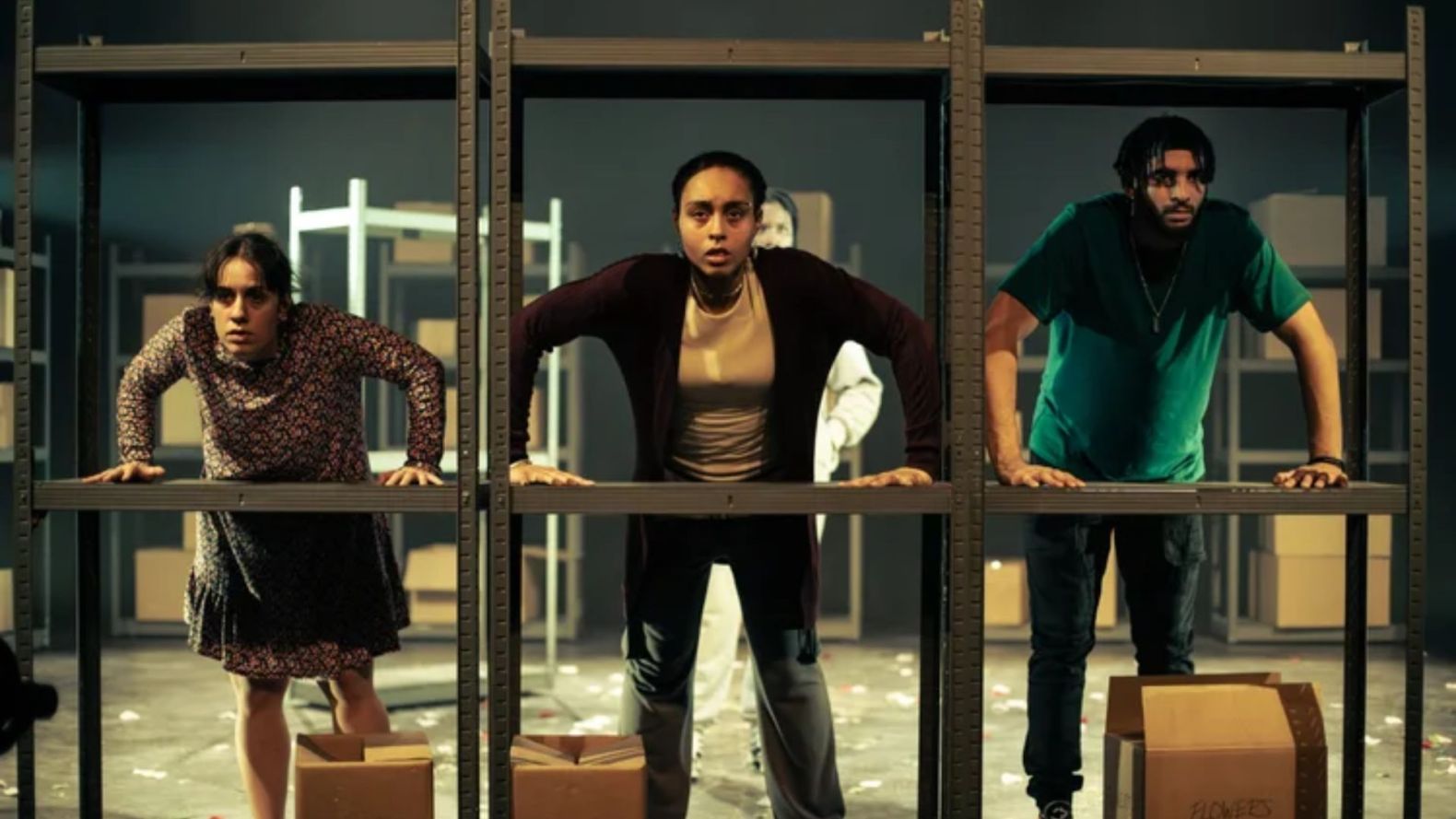 three performers in a row, their hands flat on the shelf of a metal unit, as though each of them is looking out of a window