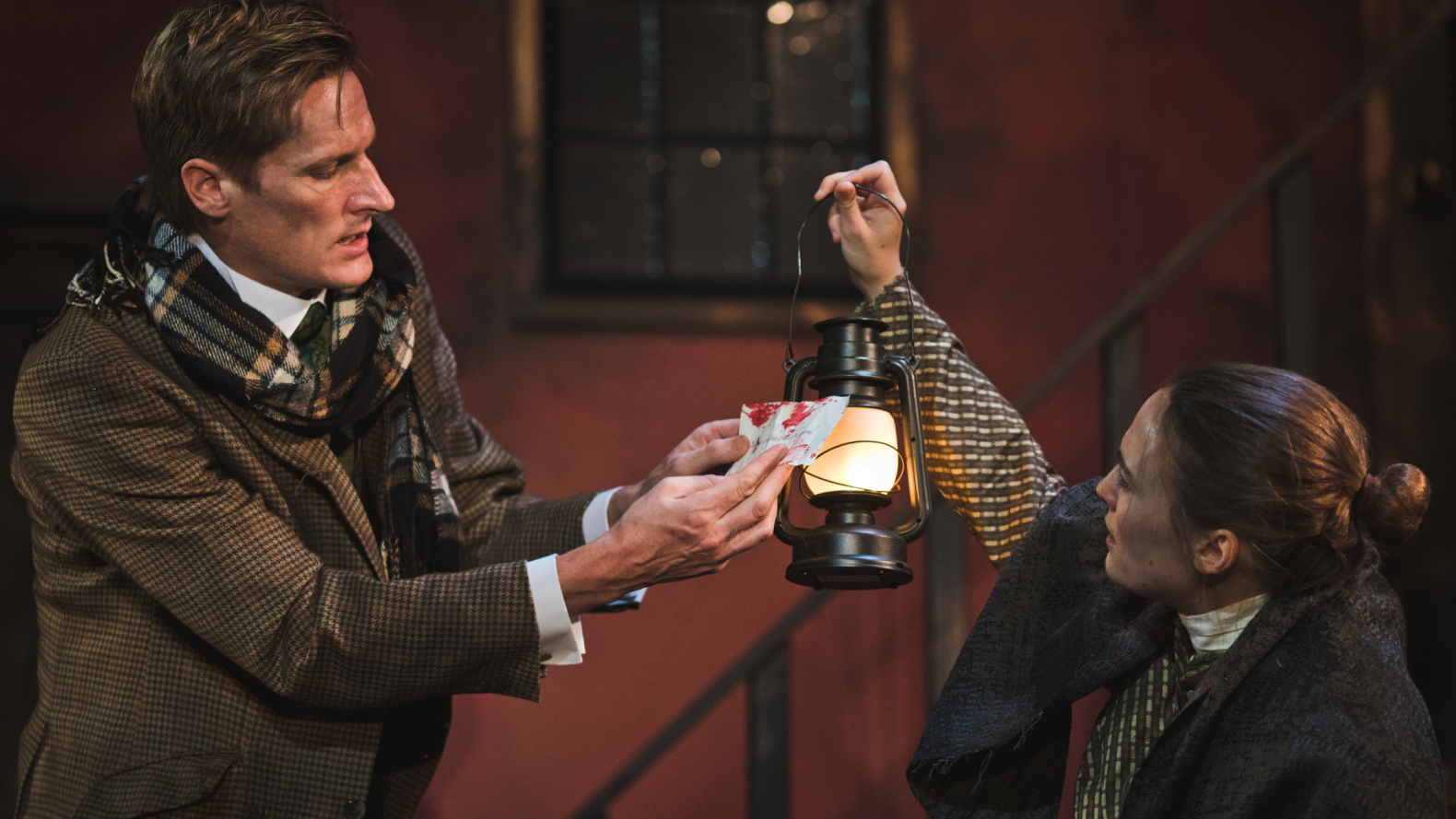 A man and a woman face each other. The woman holds up a lantern to what the man is holding: a note that is seemingly stained with blood.