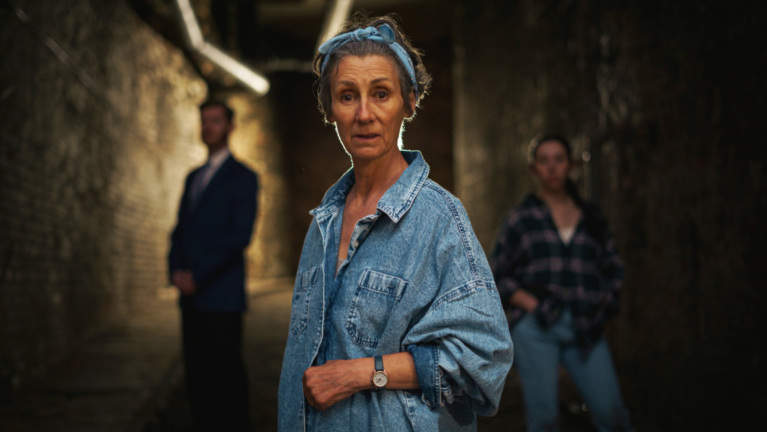 A woman in her late middle-ages. She is wearing a blue bandana on her head, tied in a bow and a oversized blue jeans shirt.