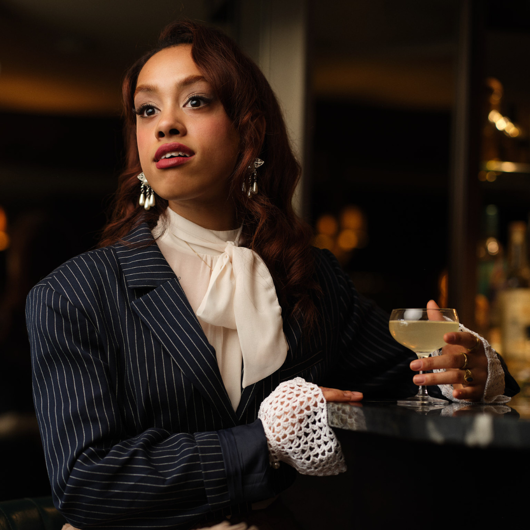 Sybil played by Hope Yolanda: Sitting at the bar, hand around a margarita. She is wearing a pin stripe jacket, with a cream blouse with a bow. 