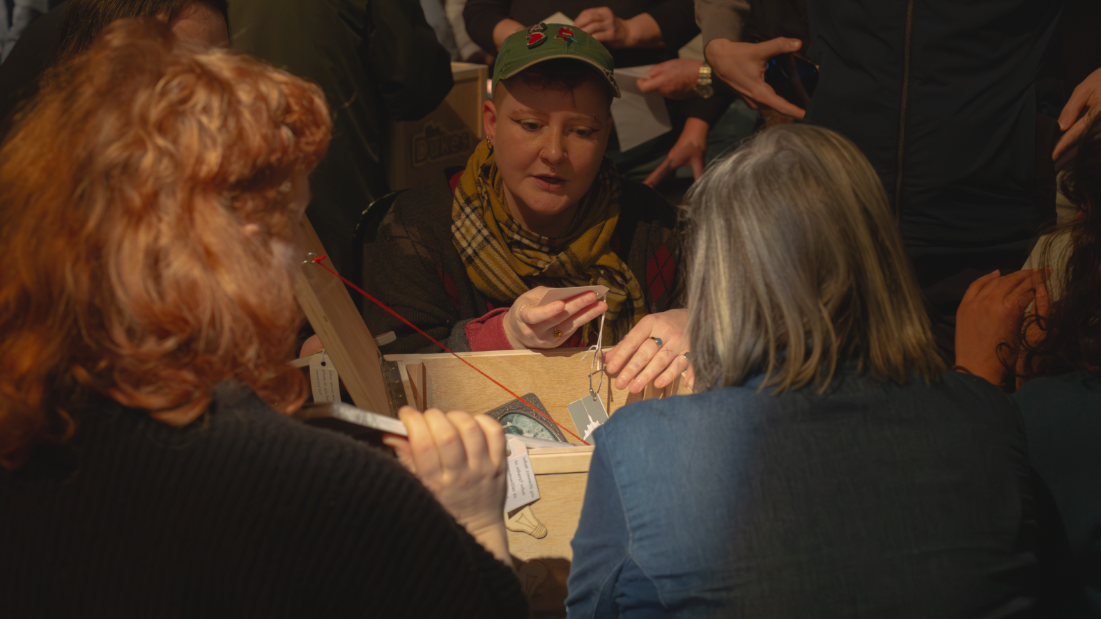 A close up of a Queer by Gum member reading a tag attached to an item from the wooden box in front of them. Two members are sitting across from them, looking inside the box. 