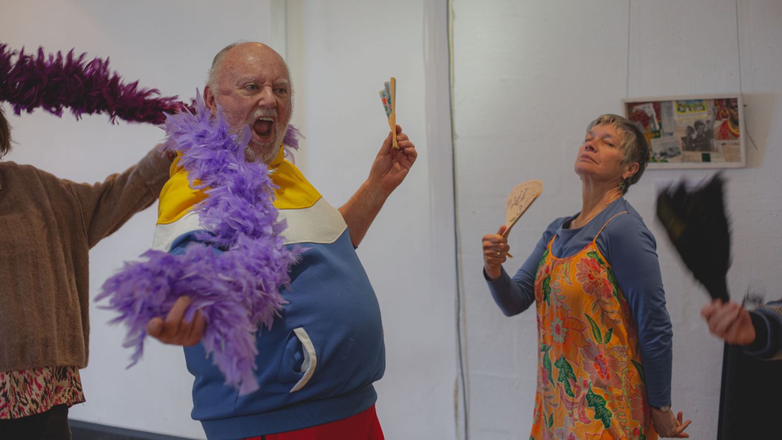 Two Prime Time members posing. One member is wearing a purple feather boa and holding a fan, and the other member is fanning themselves.