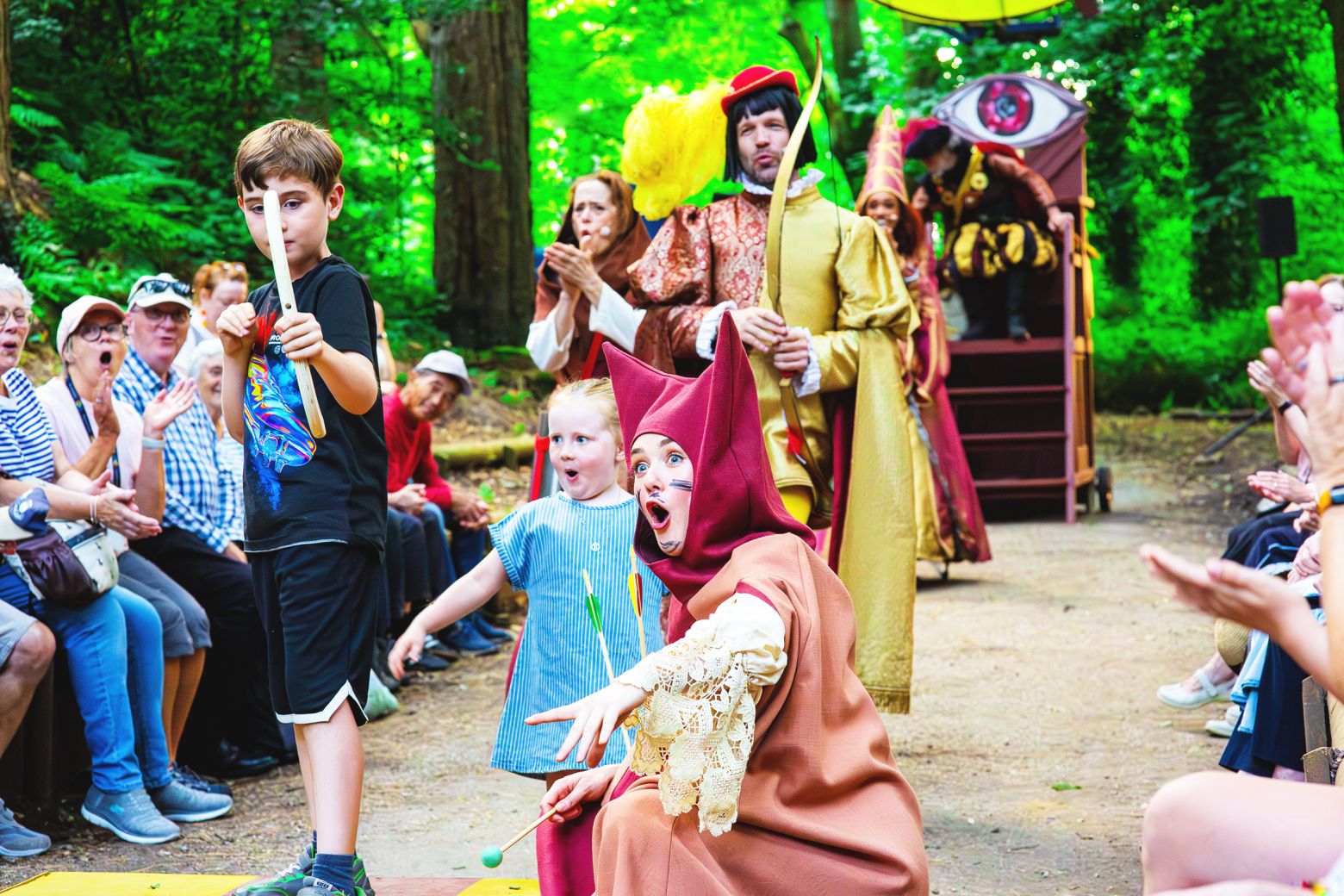 Eve Pereira, as Jen, helps some children from the audience with an archery competition in an interactive scene. Other cast members and audience can be seen in the background.
