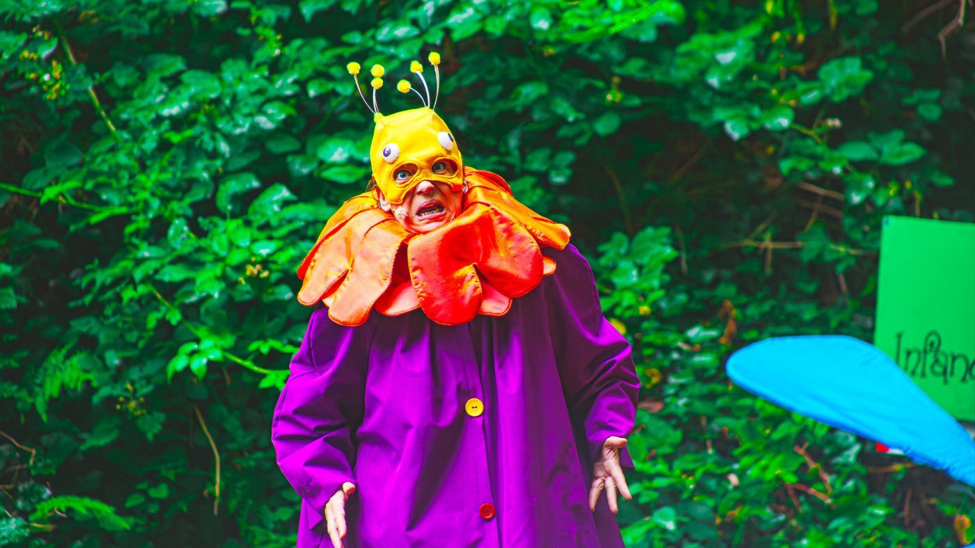A close up of a Munchkin (Helen Longworth), wearing a purple raincoat with a yellow and red button, a orange flower shaped collar, and a yellow mask, with big bold eyes and pompoms. The Wizard of Oz by the Dukes, photos by Gabi Dawkins.