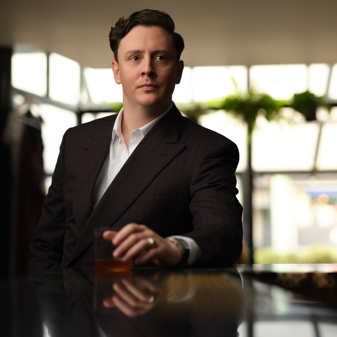 Victor played by Jack Elliot: standing at the bar, with hand over a glass of whiskey. He is wearing a black suit and white shirt. 