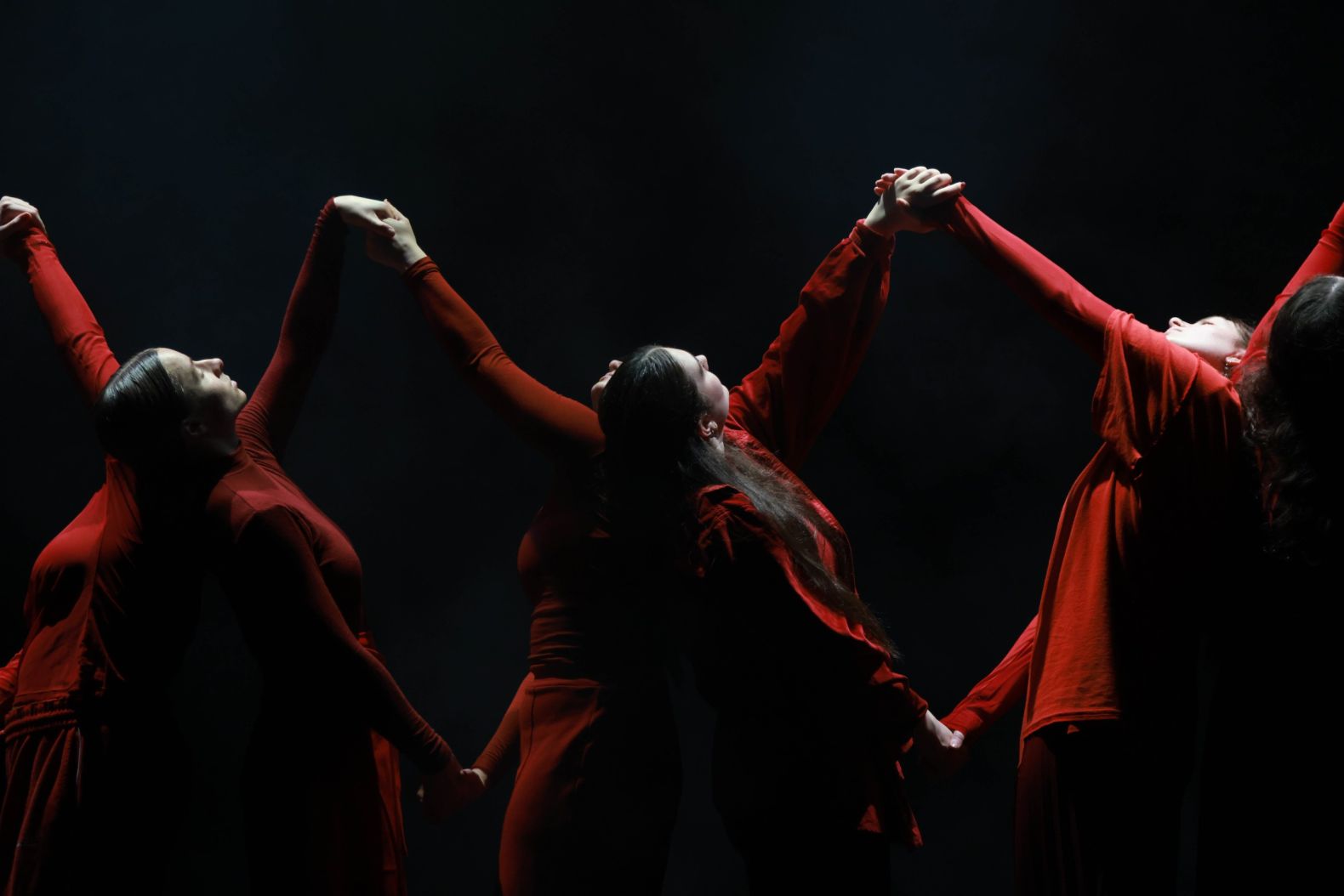 Six dancers dressed in red lean back to back, resting their heads on each others shoulders. Each left hand holds the dancer opposite's right hand, and they reach up and down alternately, forming a double helix type formation.