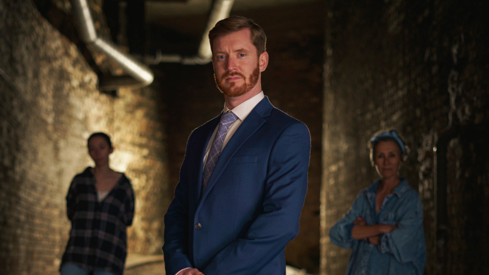A man in his 30s, with ginger hair and beard. He is wearing a navy suit, white shirt and a purple tie.