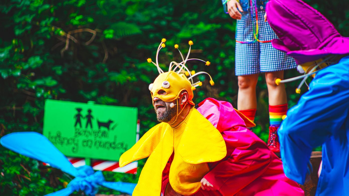 A close up of a Munchkin (Gareth Ganley), wearing a pink raincoat, a yellow flower shaped collar, and a yellow mask, with big bold eyes and pompoms. The Wizard of Oz by the Dukes, photos by Gabi Dawkins.