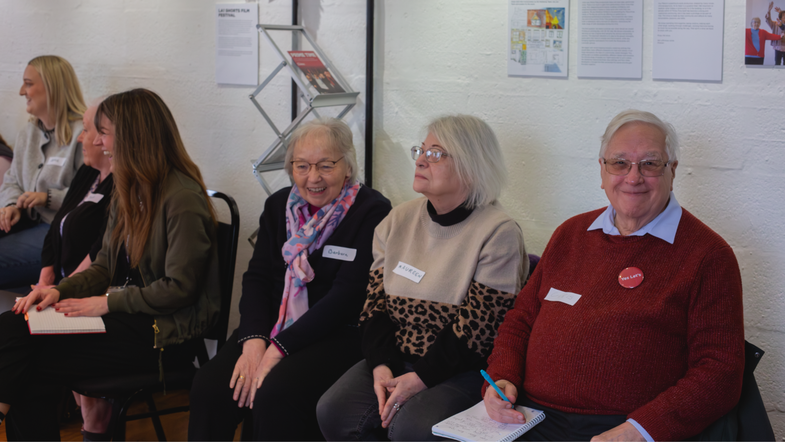 Three Prime Time members as well as other people sitting on chairs along the wall, laughing