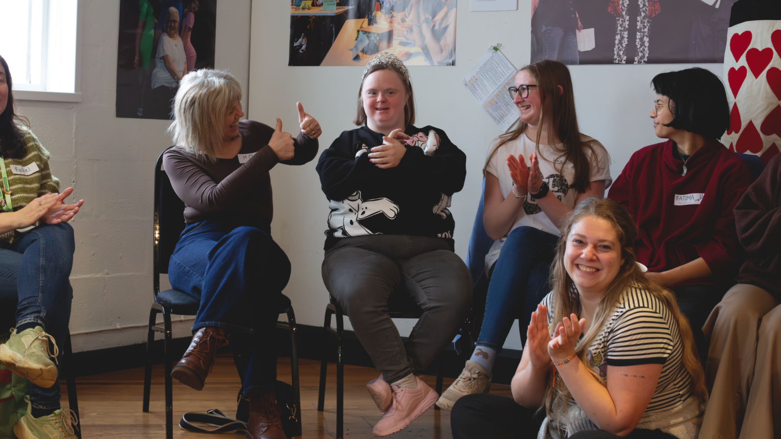 A group sitting side-by-side against the wall, with one member sitting on the floor. They are all clapping while the leader from Unique Kidz is giving two thumbs up to a member of Unique Kidz group.