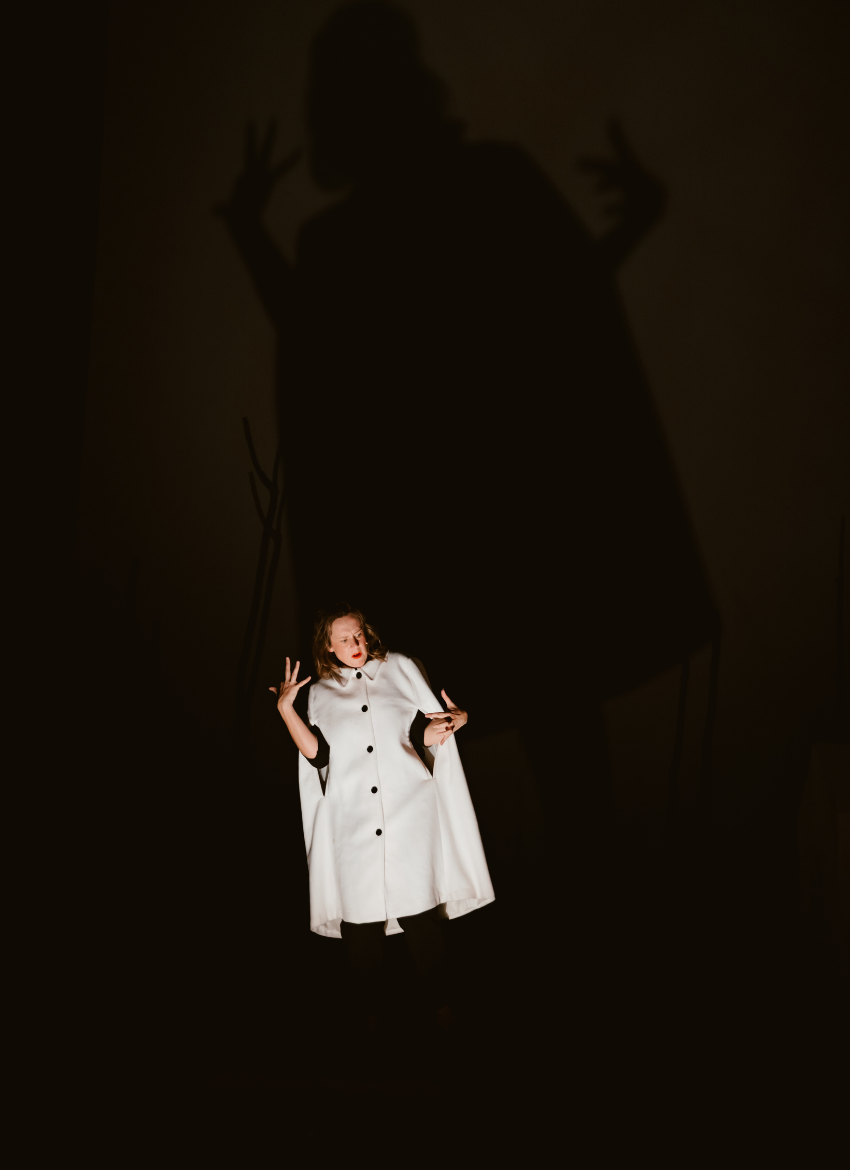 Bryony Kimmings performing in Bog Witch. Bryony is dressed in white, with a huge shadow behind her on the dark background.