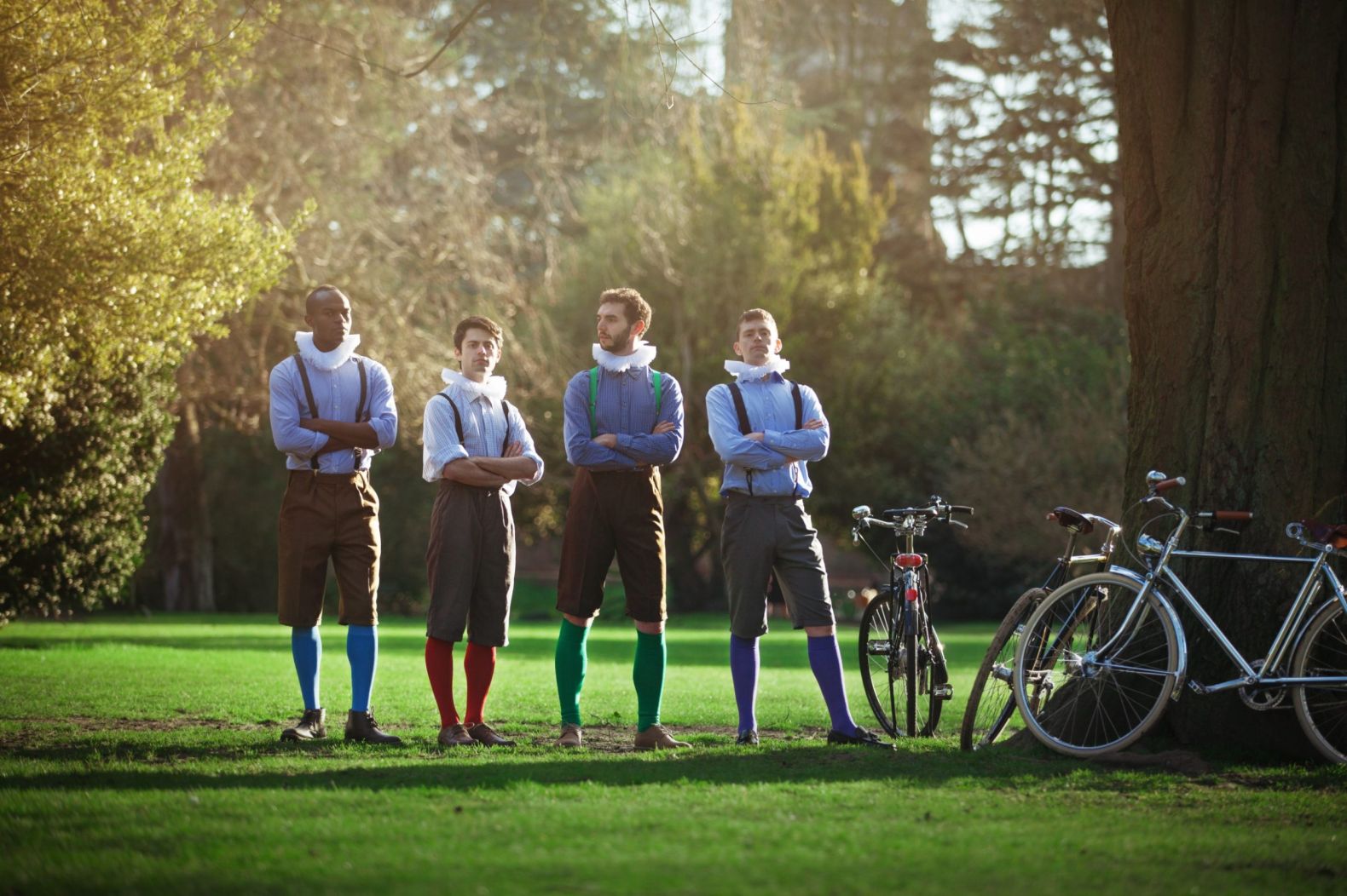 The Handlebards company stood in a park with their bikes leaning up against a tree.  They are wearing britches and ruffs