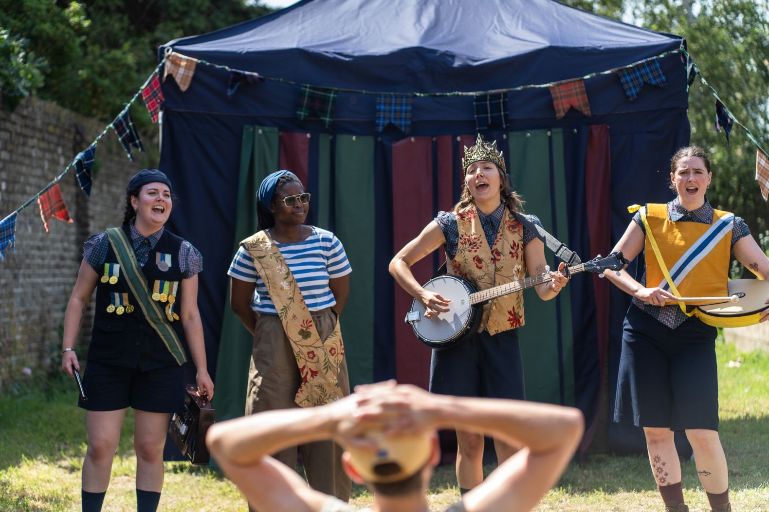 Four cast members performing outdoors in front of a tent, some playing instruments.