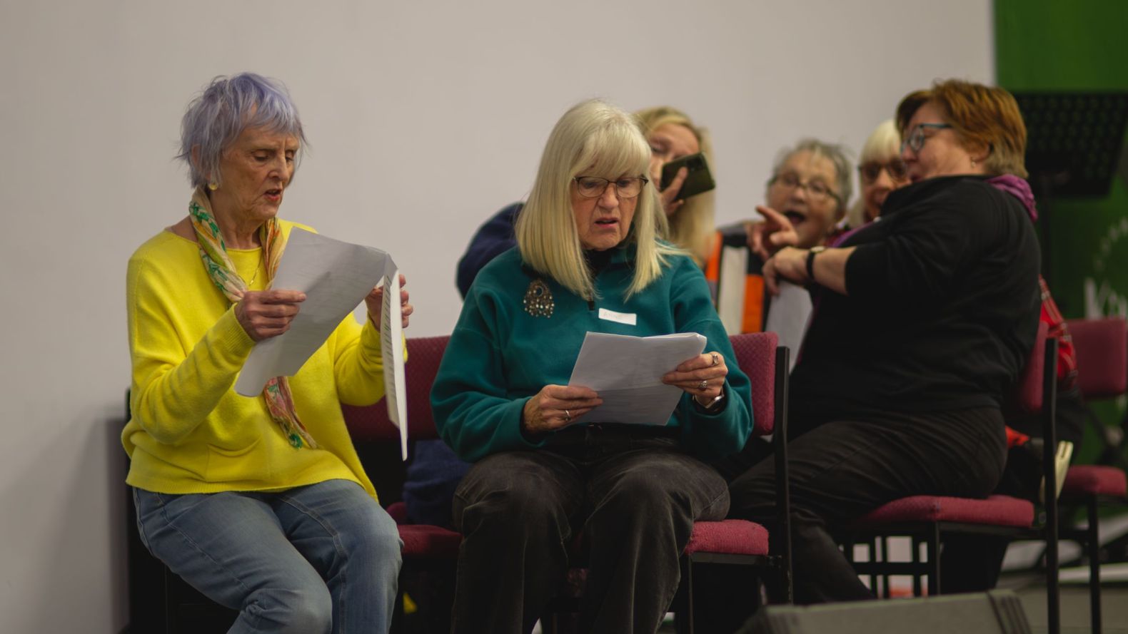 Two Prime Time Members reading of a script while four other members are sat behind them, pretending to be watching something on a phone