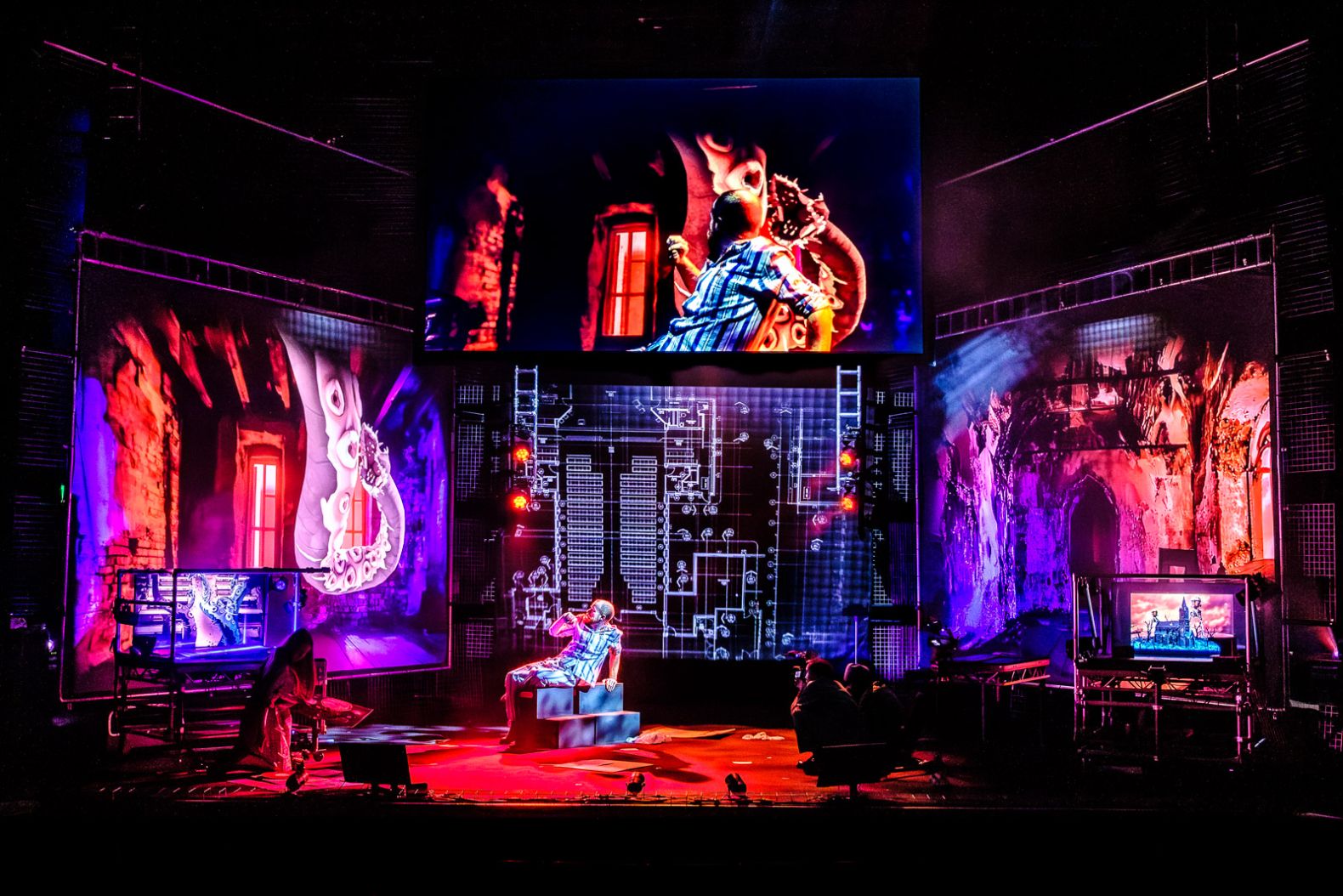 The set of War of the Worlds: Three screens surrounding stage and one hanging above, with projections, 4 actors on stage, 2 are holding camera's. On the screens you can see one actor terrified of a massive tentacle.