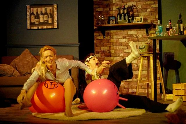 2 actors in a living room setting, on Space Hoppers.  They are both laughing and one has fallen off