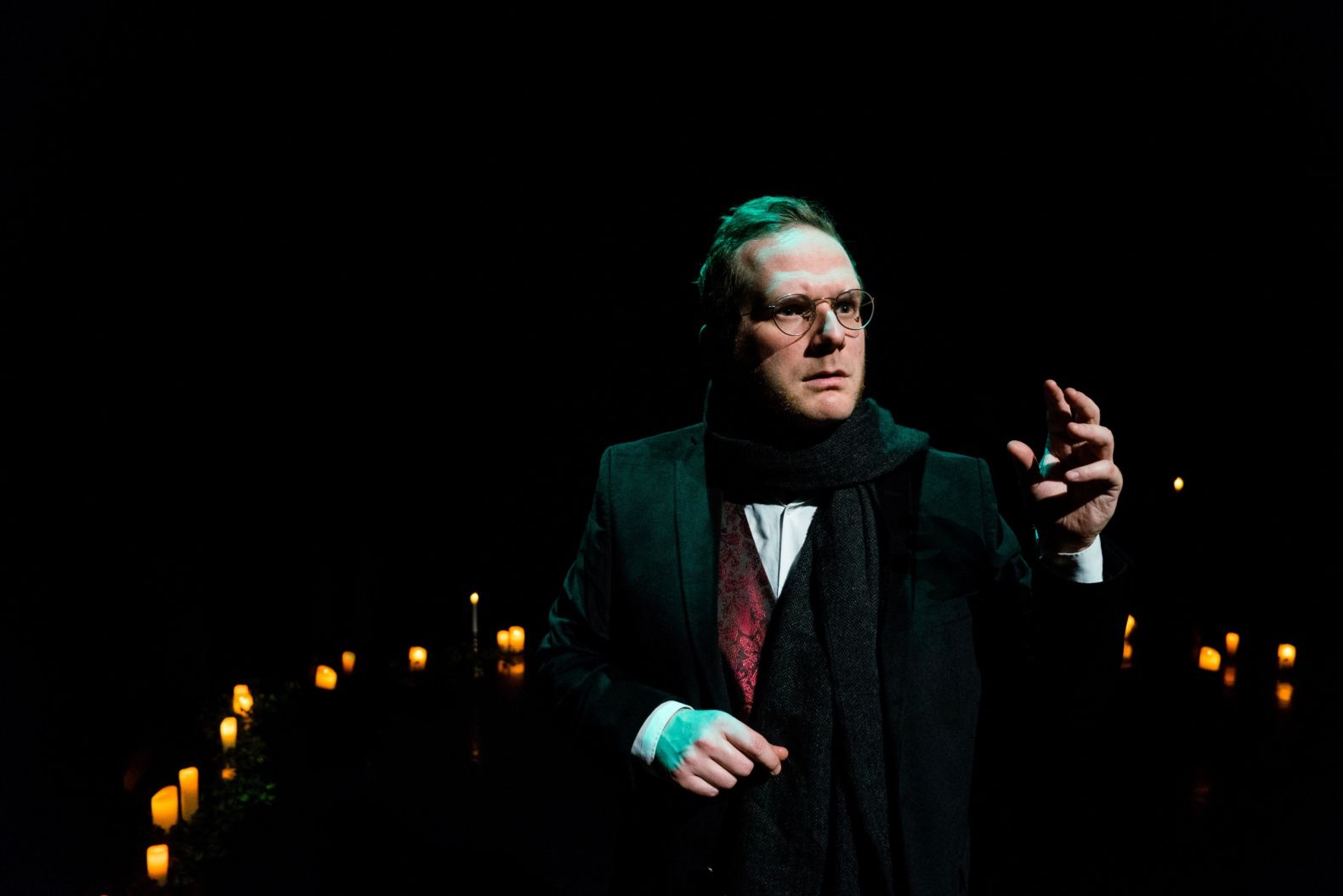 actor dressed in Victorian attire, stood surrounded by candles on the floor.  He looks as though he's heard or seen something of concern