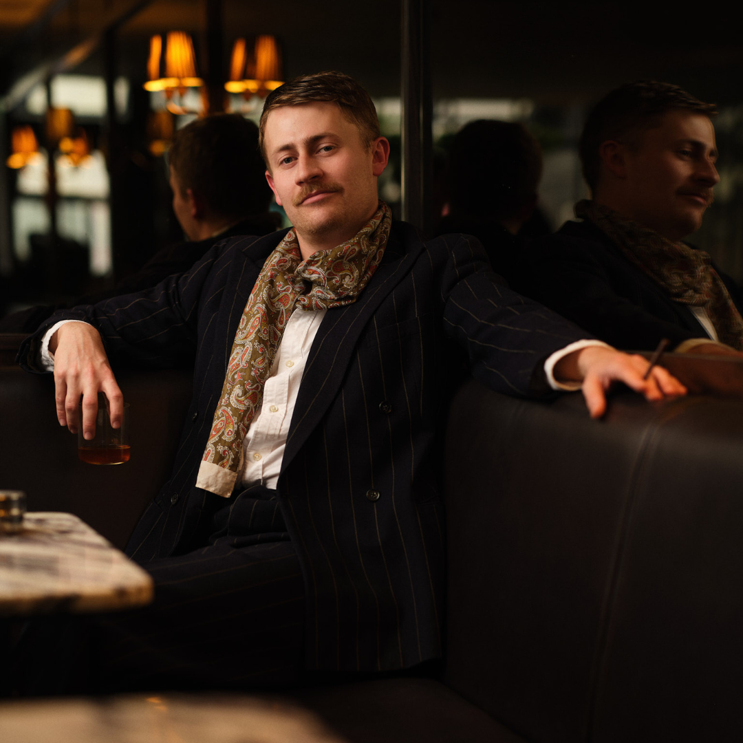 Elyot played by Charlie Noble: Sitting on a sofa with one arm resting along the top, with a cigarette between his fingers and a glass of whiskey in his other hand. 