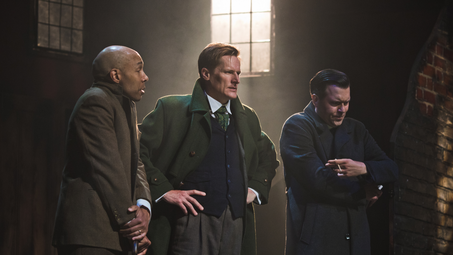 Three men stand in a line, the first has his arms crossed as if he is cold. The second, has his hands on his hips and a look on his face that suggests he is deep in thought. The third holds a cigarette whilst he folds his unoccupied arm cross his chest.