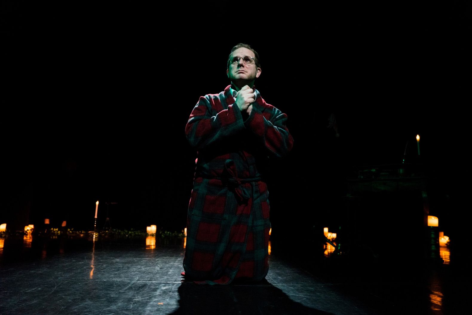 Actor dressed in a dressing gown, kneeling on the floor.  His hands clasped together as if in prayer