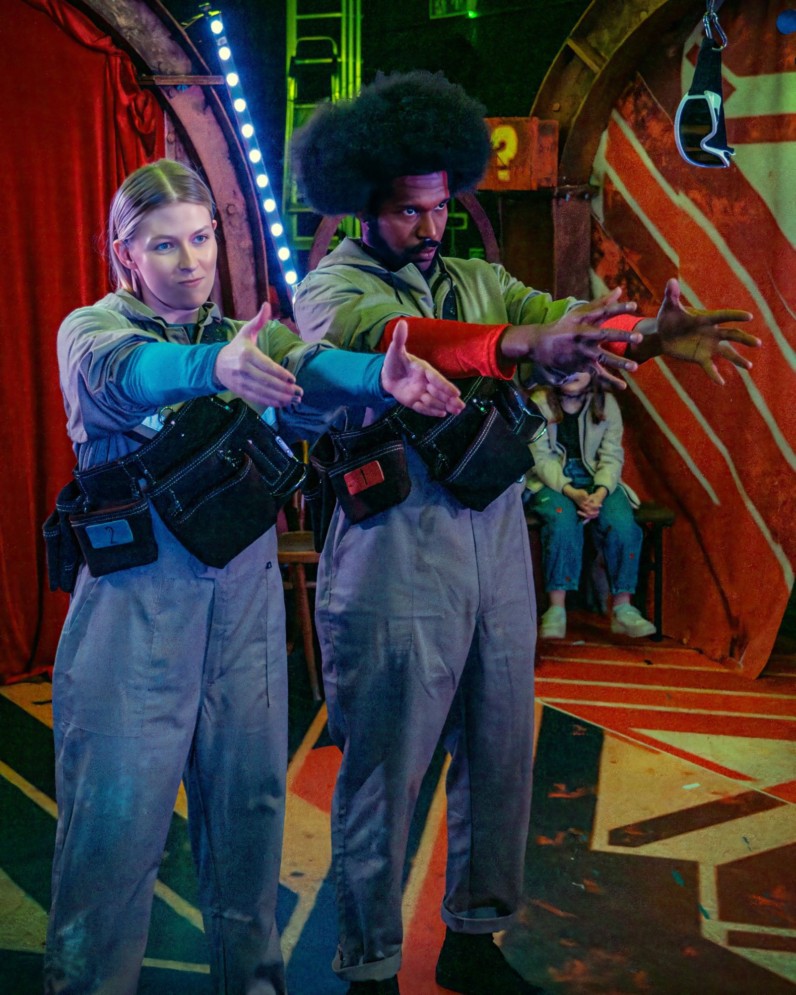 2 actors, dressed in boiler suits, sood side-by-side with their arms out-stretched in front of them