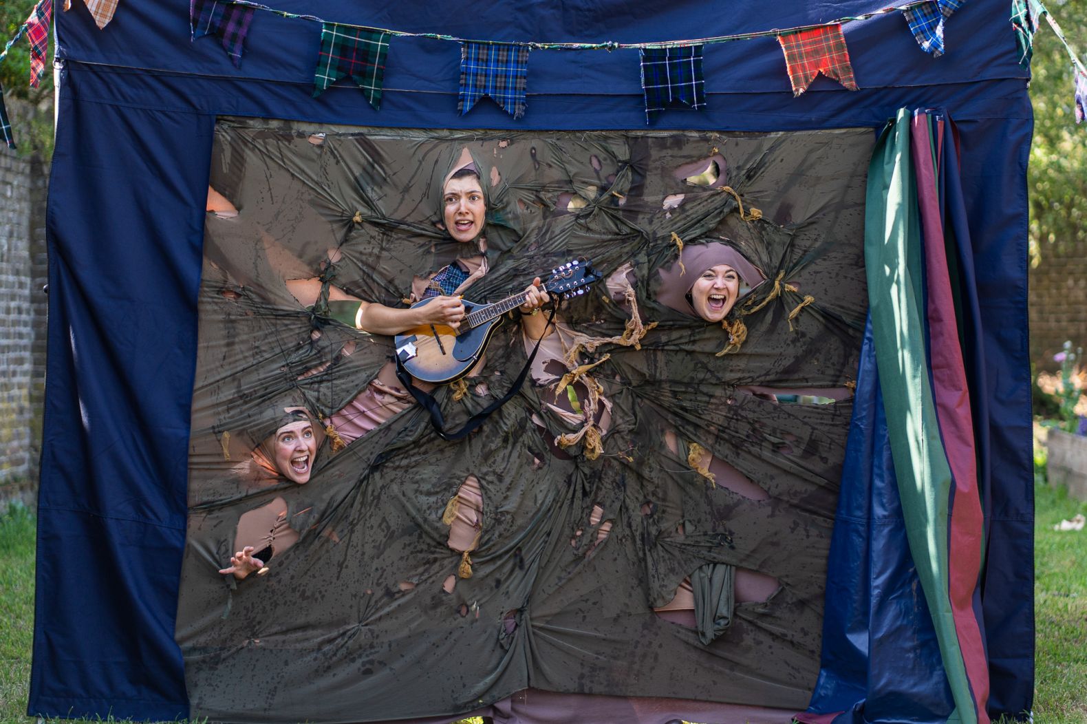 The Handlebards team performing outdoors. Three faces can be seen poking through a large fabric construction. 
