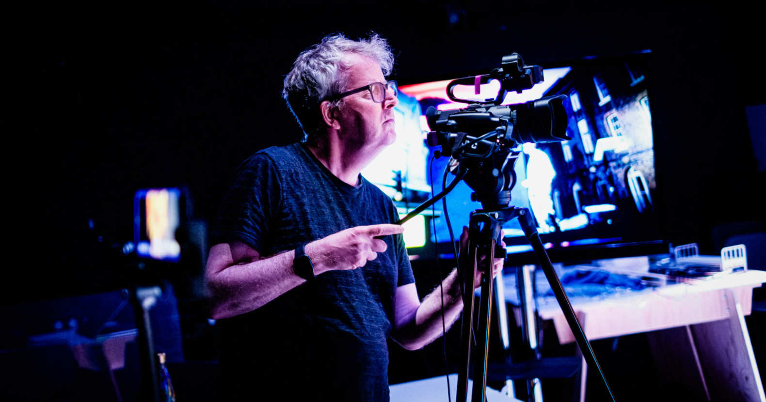 A man in his 50s, wearing a black top, and watch, operating a camera on a tripod.