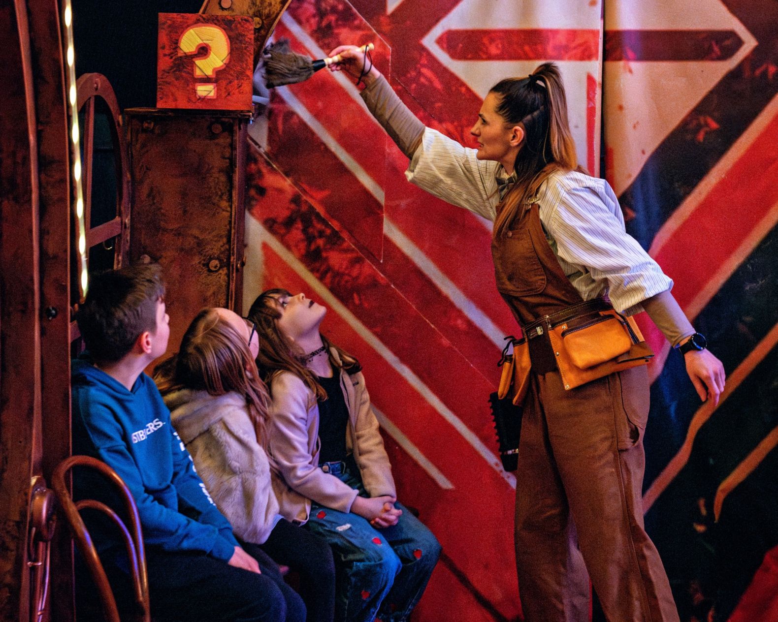an actor, dressed in dungarees and a large tool-belt, dusts part of the set above children sat in the audience