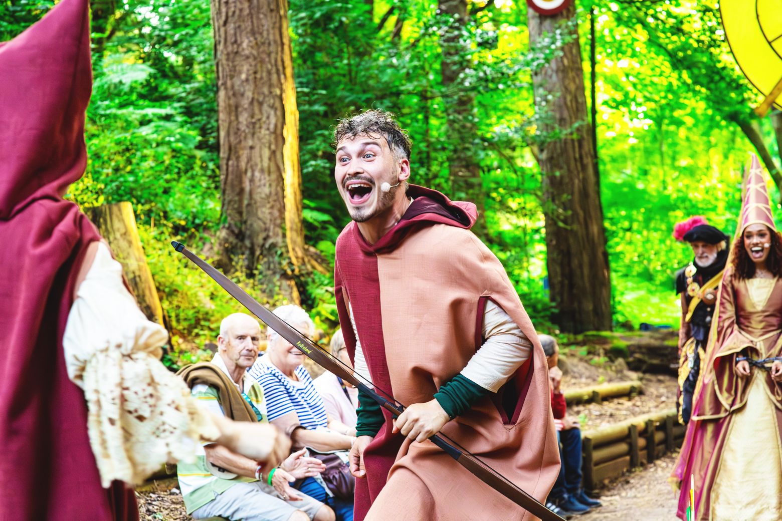 Joe Parker as Robin in the park, holding a bow-and-arrow and laughing.