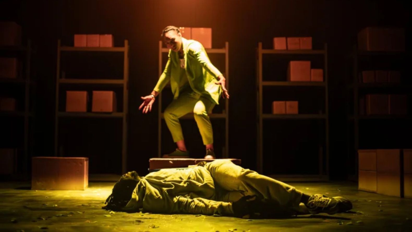 A performer lies on the floor as another looms over them, stood half squatting on a box